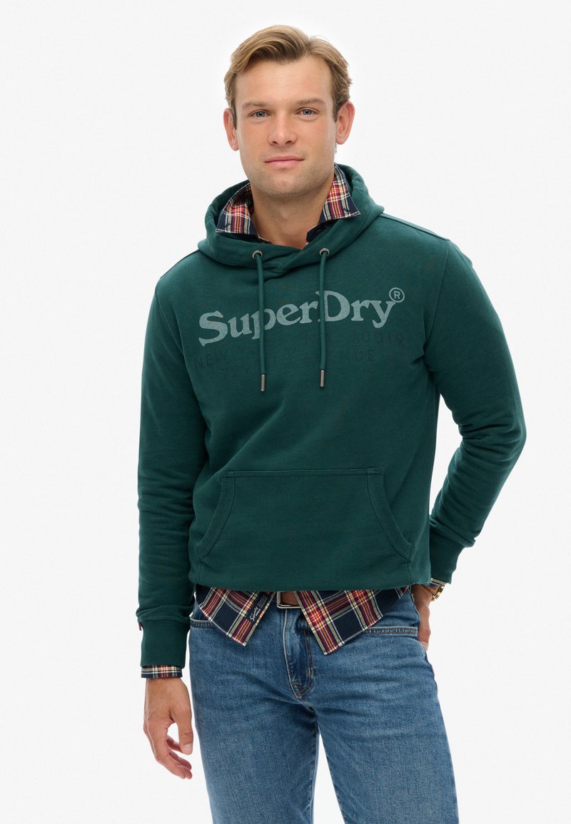 Zielona bluza z kapturem, sznurkami i kieszenią z przodu, z dużym szarym logo "SuperDry". Noszona na koszuli w kratę. Casualowy krój i faktura.