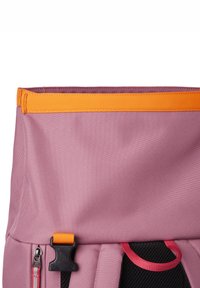 Primer plano de una mochila morada con una correa naranja, hebilla negra y lazo rojo, mostrando la textura de la tela y los detalles del cierre.