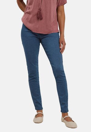 Kvinde iført blå skinny jeans, en støvet rosa bluse med frynser og beige Mary Jane-sko, stående mod en ensfarvet baggrund.