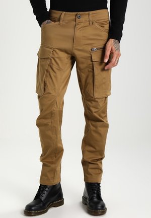Man wearing brown cargo pants with side pockets and black lace-up boots, standing against a plain background.