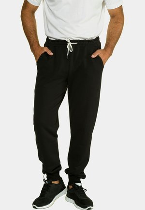 Pantalones de chándal negros de tela suave con cintura elástica y cordón; cuentan con bolsillos laterales y puños ajustados. Combinados con zapatillas negras.