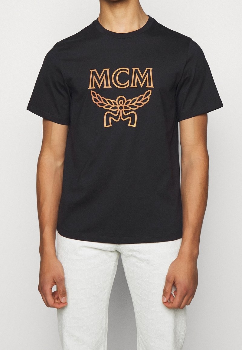 Camiseta de algodón negra con el logo "MCM" en naranja y diseño decorativo de alas. Mangas cortas y un cuello redondo clásico.