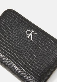 Portofel negru din piele ecologică cu un model texturat asemănător crocodilului. Dispune de logo-ul argintiu "CK" și o închidere cu fermoar pe margine.