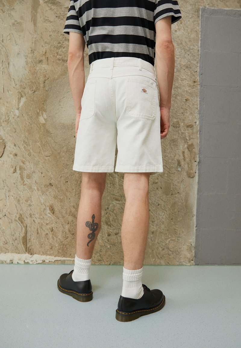 Hvide denim shorts med to baglommer, parret med sorte sko og hvide sokker. Synlig slange tatovering på underbenet. Stripet skjorte ikke synlig.