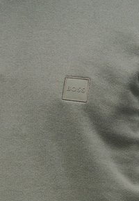 Φωτεινό πράσινο πλεκτό πουλόβερ με τετράγωνο λογότυπο patch με το κείμενο "BOSS" στην κάτω αριστερή γωνία, αναδεικνύοντας την υφή του υλικού.