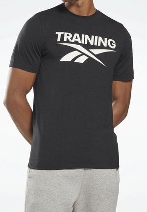 Uomo che indossa una maglietta nera con la scritta "TRAINING" e un logo stilizzato bianco, abbinata a pantaloni della tuta grigio chiaro, in piedi con le mani dietro la schiena.