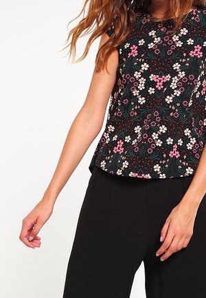 Femme portant un haut noir à manches courtes avec des motifs floraux roses et blancs, associé à un pantalon noir taille haute.