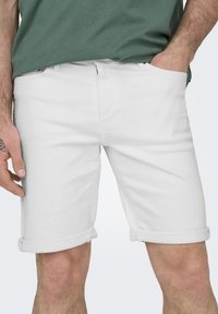 Shorts de mezclilla blanca con dobladillos enrollados, que presentan un corte recto y bolsillos frontales. Llevados con una camiseta verde, destacando el diseño de los shorts.