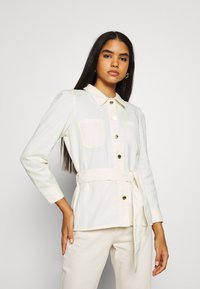 Camicia a maniche lunghe con bottoni in tessuto crema chiaro, con colletto, due tasche sul petto e una cintura in vita, dotata di bottoni dorati.