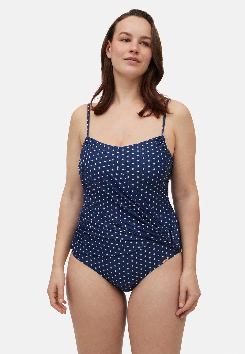 Traje de baño de lunares azul marino con tirantes de espagueti ajustables, que presenta un diseño envolvente y una textura suave. Detalle de acento plateado en el costado.