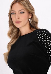 Zwarte top van zacht stof, met glanzende rhinestones op één schouder en een elegante halslijn. Model heeft lang golvend haar en oorbellen.