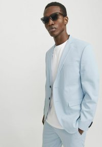 Hellblaues Anzug mit taillierter Passform, bestehend aus einem einreihigen Jacket und passender Hose, getragen über einem weißen T-Shirt. Schwarze Sonnenbrille.