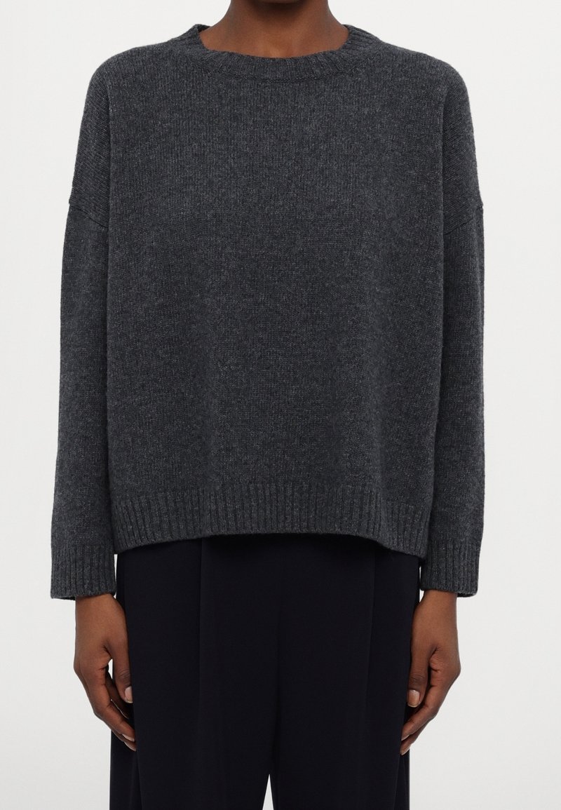 Pull en maille gris foncé à manches longues avec un col rond, porté avec un pantalon noir, visible des épaules aux hanches sur un fond uni.