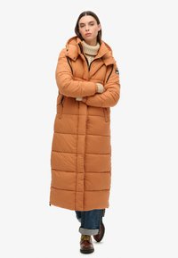 Lang, gewatteerde jas in warm oranje, met een capuchon, ritssluiting aan de voorkant, zijzakken en geribde boorden. Textuur stof met een horizontaal gestikt patroon.