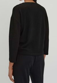 Černý pletený svetr s vroubkovanou strukturou, širokými rukávy a kulatým výstřihem, nošený s tmavými kalhotami, ukazující pohled zezadu.