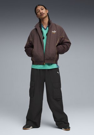 Veste bomber marron avec motif texturé et accents jaunes, chemise verte en couches, pantalon cargo noir et baskets noires.