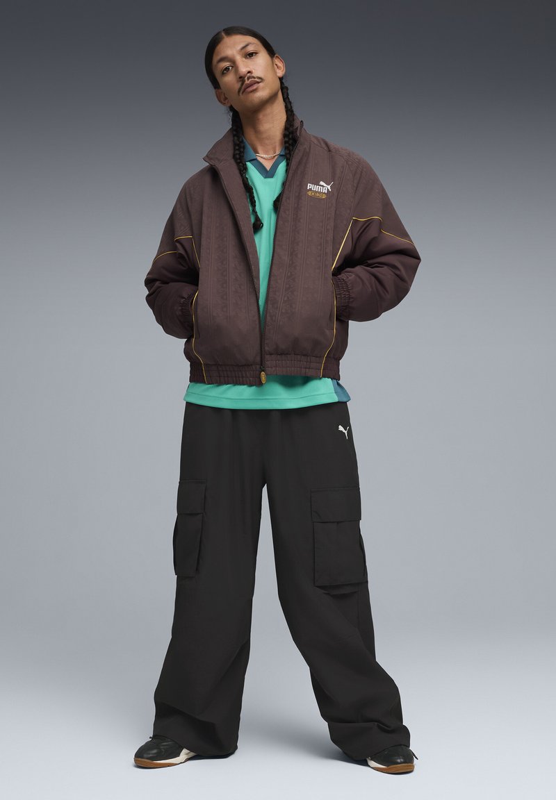 Barna bomber dzseki texturált mintával és sárga díszítéssel, zöld réteges ing, fekete cargo nadrág és fekete sportcipő.