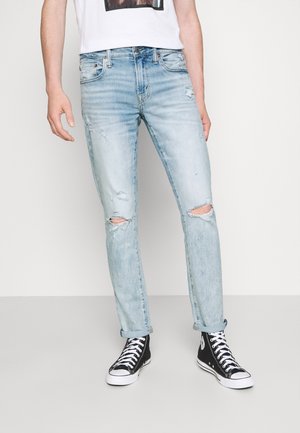 Mann trägt hellblaue, zerrissene Jeans, die an den Knöcheln aufgerollt sind, kombiniert mit schwarzen High-Top-Sneakern und einem weißen T-Shirt mit Grafik.