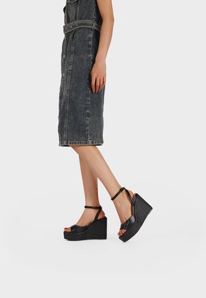 Donna con sandali con zeppa neri con cinturini alla caviglia e una gonna di jeans scura a vita media con bottoni, in piedi di lato su sfondo bianco.