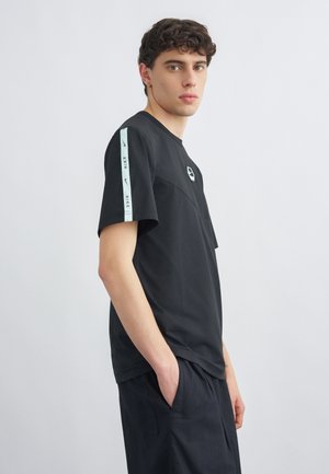 Junger Mann mit lockigem Haar, der ein schwarzes Nike-T-Shirt und schwarze Hosen trägt, seitlich stehend mit den Händen in den Taschen vor einfarbigem Hintergrund.