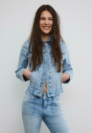 Jeune femme souriante aux longs cheveux bruns, portant une veste en denim bleu clair et un jean, les mains dans les poches de la veste, sur un fond uni.