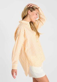 Femme souriante les yeux fermés, portant une chemise oversize rayée jaune et blanche et une jupe blanche, la main reposant sur la tête.