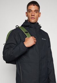Veste imperméable noire avec capuche, dotée d'une fermeture éclair à l'avant, de poignets élastiques et d'un logo. Le mannequin porte un sac à dos avec une bandoulière verte.