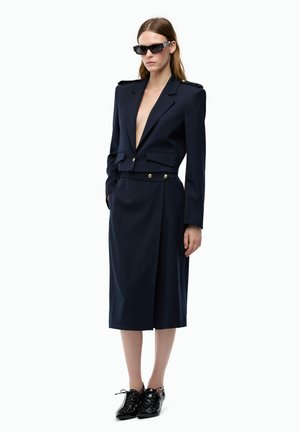 Donna che indossa un blazer sartoriale blu scuro con bottoni dorati, gonna midi abbinata, scarpe nere stringate e occhiali da sole neri rettangolari.
