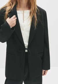 Femme portant un blazer noir cintré à la taille par-dessus une chemise blanche à boutons, avec de longs cheveux blonds, montrée des épaules aux hanches.