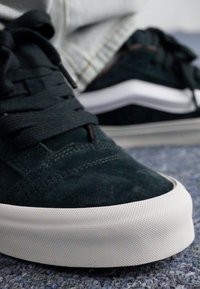 Baskets en suede noir avec semelle en caoutchouc blanc, présentant un design profil bas, un bout rond et des lacets texturés. Détails de couture subtils visibles.