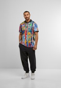 Bărbat în picioare, îmbrăcat cu o cămașă scurtă cu un model abstract colorat, pantaloni de jogging negri și adidași albi, pe un fundal simplu.