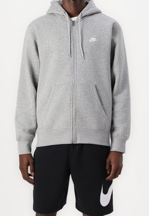 Homme portant un sweat à capuche zippé gris clair Nike avec capuche et poches avant, associé à un short Nike noir avec un grand logo.
