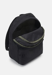 Mochila de tela negra con una solapa de forro verde, apertura con cremallera y bolsillo exterior; cuenta con correas de hombro acolchadas y una estructura resistente.