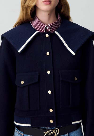 Chaqueta recortada de lana azul marino con gran cuello acanalado, detalles en blanco, botones dorados, doble bolsillo frontal y un cinturón negro sobre unos vaqueros de lavado claro.