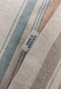 Primo piano di un tessuto con righe intrecciate nei toni del blu, beige e marrone, con un'etichetta che recita "COLOURS & SONS."