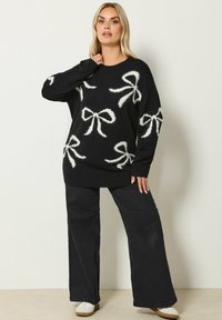 Czarny sweter z białym, puszystym wzorem w kokardki, szerokie rękawy, ściągacze u dołu i przy rękawach. W zestawie z czarnymi spodniami o szerokich nogawkach i jasnym obuwiem.