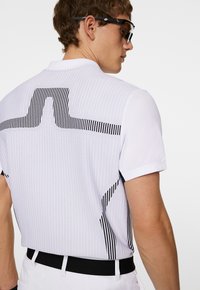 Vit polo skjorta med vertikal ribbad textur, med en svart randig design på baksidan. Svart midjeband fulländar den sportiga looken.