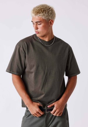 Jeune homme aux cheveux blonds courts portant un t-shirt sombre oversize et un collier chaîne, regardant vers le bas et ajustant sa ceinture.