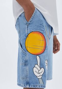 Shorts de mezclilla en azul claro con estampado gráfico. Presentan un sol amarillo brillante y una mano de dibujos animados señalando. Diseño clásico de cinco bolsillos.
