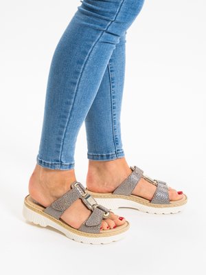 Beine in schlanken blauen Jeans, die graue Sandalen mit offenen Zehen, weißen Sohlen und metallischen Schnallendetails tragen, auf einem weißen Hintergrund stehend.