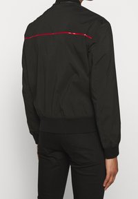 Giacca bomber nera con una texture liscia, caratterizzata da una striscia riflettente rossa sulla schiena e polsini e orlo a coste.