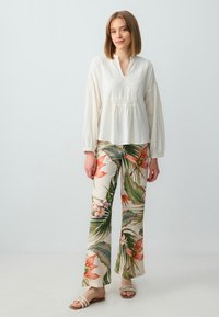 Blusa bianca a maniche lunghe con dettagli ricamati sul davanti, abbinata a pantaloni flare con fantasia floreale in tonalità verdi, rosse e rosa.