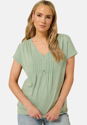 Pitkähiuksinen nainen, jolla on vaaleanvihreä, lyhythihainen pusero, jossa on napit ja laskokset, yhdistettynä vaaleansinisiin farkkuihin.