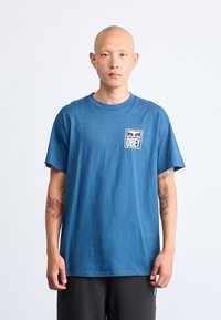 Blå bomull T-skjorte med korte ermer og rund hals. Har en rektangulær hvit lapp med ordet "OBEY" og grafisk design.