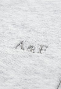 Lys grått stoff med en myk tekstur som har broderte sølvfargede bokstaver "A&F" i et uformelt skrifttype. Mønsteret er subtilt og ensartet.
