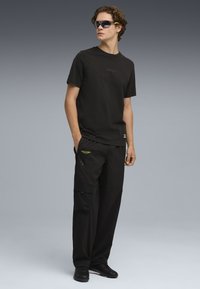Schwarzes Baumwoll-T-Shirt mit dezentem Logo, kombiniert mit schwarzen Cargo-Hosen mit Taschen und grüner Stickerei, abgerundet durch schwarze Schuhe und eine Sonnenbrille.