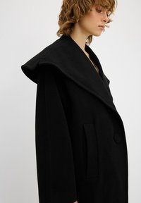 Cappotto oversize nero con un grande colletto ripiegato. Realizzato in un tessuto morbido, presenta una tasca laterale e una chiusura con un singolo bottone rotondo.