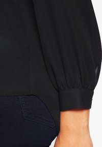 Zwarte blouse met gepofte mouwen en een gladde textuur, voorzien van een snug passende manchette en een lichte glans. Gecombineerd met donkerblauwe denim.