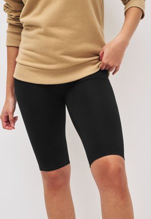 Personne portant des leggings noirs longueur genou et un haut beige à manches longues, tenant l'ourlet du haut avec une main.