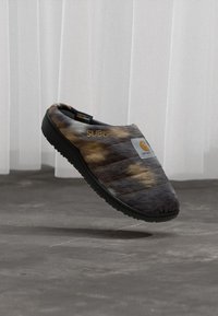 Braun und schwarz gefärbte Slip-on-Schuhe mit schwarzer Sohle, mit "SUBU" und Carhartt-Logopatches, über einem grauen Boden vor weißen Vorhängen schwebend.